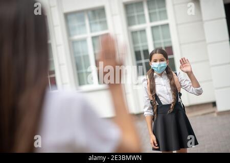 Schulmädchen in schützende Maske sagen Auf Wiedersehen zu Mutter Stockfoto