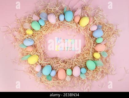 Bunte Ostereier und Federn in Pastellfarben liegen auf Spänen in Form eines Rahmens auf rosa Farbe. Die Inschrift Frohe Ostern im Zentrum Stockfoto