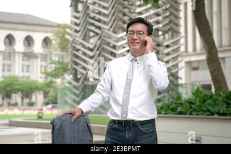 Asiatischer Geschäftsmann lächelnd und am Telefon sprechend.beim Halten auf Jacke. Außenumgebung. Stockfoto