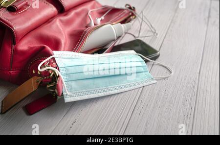 Frau rote Handtasche mit Gesichtsmaske, Handdesinfektionsmittel, Ohrhörer und Smartphone auf Holztisch. Stockfoto