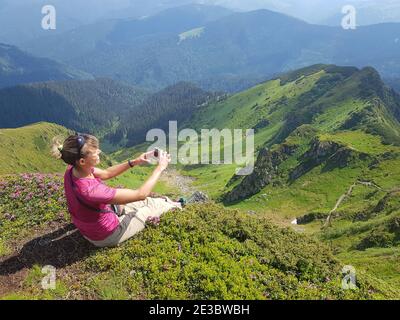 Mädchen sitzt auf einer Wiese mit rosa Blumen und macht Bilder von Bergen per Smartphone. Maramures Region der Karpaten. Stockfoto