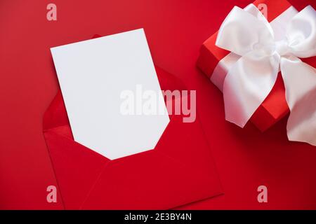 Grußkarte Mockup, weiße leere Karte im Umschlag und Geschenkbox auf rotem Hintergrund. Alles Liebe zum Valentinstag oder zum Muttertag. Flach liegend, Draufsicht. Stockfoto