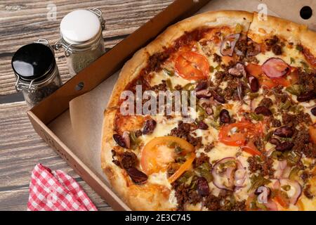 Draufsicht auf eine frisch gekochte Pizza in einer Pappschachtel, Pizzabesser auf einer Serviette und Salz und Pfeffer auf einem Holztisch. Pizzalieferung. Essen Sie zu Hause. Stockfoto