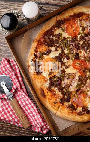 Draufsicht auf eine frisch gekochte Pizza in einer Pappschachtel, Pizzabesser auf einer Serviette und Salz und Pfeffer auf einem Holztisch. Pizzalieferung. Essen Sie zu Hause. Stockfoto