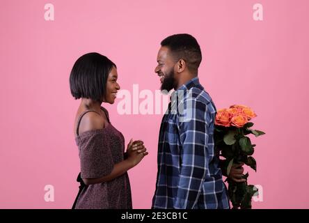 Hübsche schwarze Frau im noblen Kleid und ihr Freund versteckt Blumenstrauß hinter seinem Rücken auf rosa Studiohintergrund Stockfoto