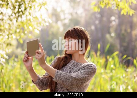 Eine schöne Frau mit langen Haaren sitzt in einem Frühlingspark im Freien, mit einem digitalen Tablet. Stockfoto