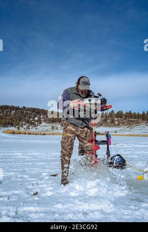 Ein Mann in der camofloge-Hose bohrt ein Loch in das schneebedeckte Eis zum Eisfischen auf dem Echo Canyon Reservoir in der Nähe von Pagosa Springs, Colorado. Stockfoto