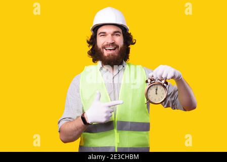 Foto des staunenden bärtigen Architekten, der Helm trägt und auf Wecker zeigt. Stockfoto