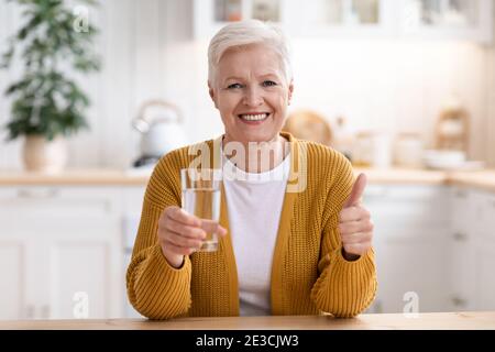 Fröhliche ältere Frau mit einem Glas Wasser, das den Daumen nach oben zeigt Stockfoto