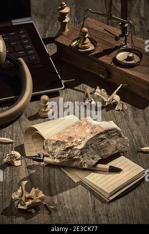 Schreibblockierung. Konzept Stillleben, Anordnung auf dunklem Holztisch. Schwerer Stein, Ziegel auf offener, leerer Blocknote. Laptop, Kopfhörer, Vintage-Waage Stockfoto