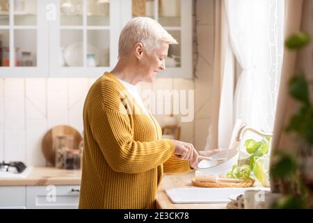 Glückliche ältere Frau, die in der Küche Geschirr wäscht Stockfoto