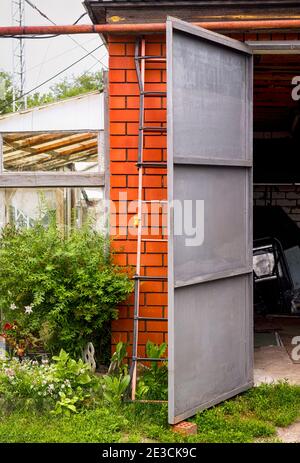 Alte Backsteingarage mit offener Tür. Es ist eine Leiter gegen die Garage gestützt. Es ist ein Gewächshaus angeschlossen. Stockfoto