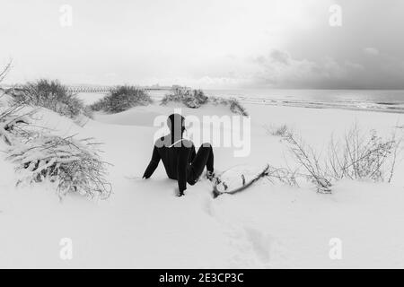 Winter und Surfer mit Surfbrett auf Schnee sitzen. Schneebedeckter Strand und Surfer im Neoprenanzug. Stockfoto