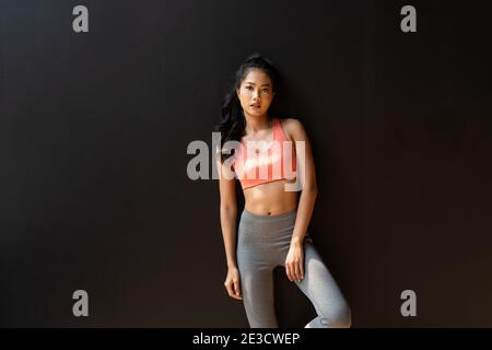 Seriöse und entschlossene Jugendliche asiatische Sportlerin, die die Kamera anschaut Beim Einstecken der Hand in die Tasche und beim Stehen vor schwarzem Hintergrund Stockfoto