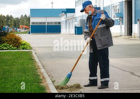 Dies ist ein älterer Hausmeister mit einem Besen in einer medizinischen Maske auf der Straße fegen das Gebiet. Stockfoto