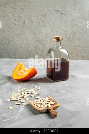 Eine Flasche Kürbiskernöl, eine Scheibe Pumpkin- und Pumpkin-Samen in einem Holzlöffel auf grauem Hintergrund, vertikales Foto mit Kopierraum Stockfoto