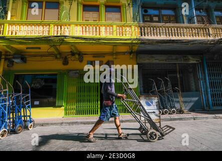 Ein Mann schiebt einen leeren Trolley entlang einer sonnenbeschienenen Straße in Chinatown, Bangkok, Thailand Stockfoto