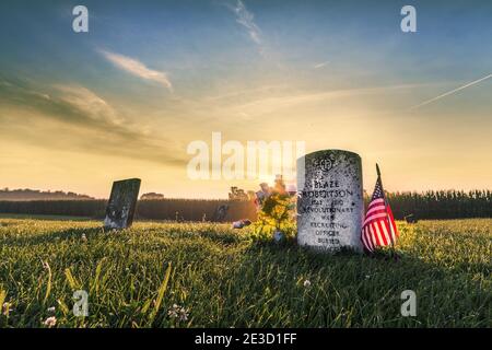 Sonnenaufgang über dem Grabstein eines revolutionären Kriegssoldaten am 4. Juli. Der Grabstein wird von einer amerikanischen Flagge und Blumen flankiert. Stockfoto