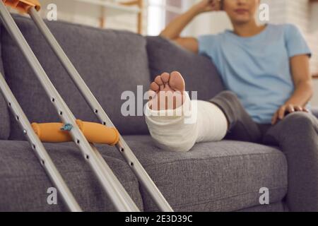 Nahaufnahme einer Krücken und eines gebrochenen Beins in einem Gipsabguss einer Frau, die auf einem Sofa sitzt und sich ausruht. Stockfoto