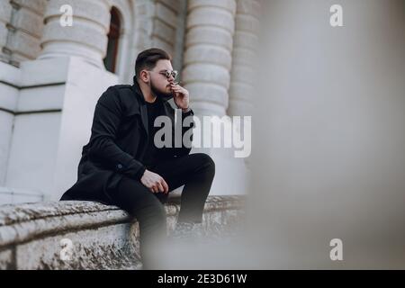 Ruhiger junger bärtiger Kerl in Sonnenbrillen rauchen, während draußen sitzen Stockfoto