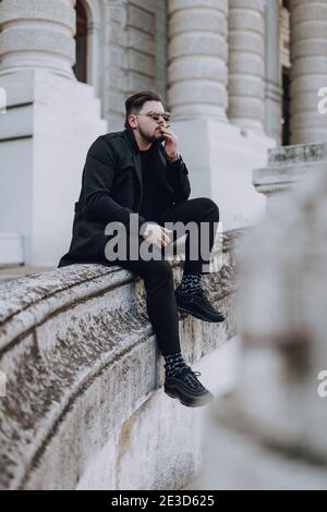 Junger Mann in Sonnenbrille sitzt in der Stadt und raucht Beim Blick vor ihn Stockfoto