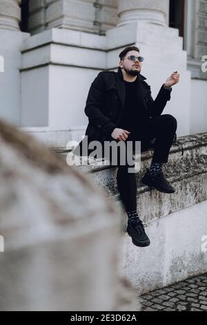 Volle Länge der stilvollen Kerl in Sonnenbrille ruht in der Stadt während des Rauchens Stockfoto