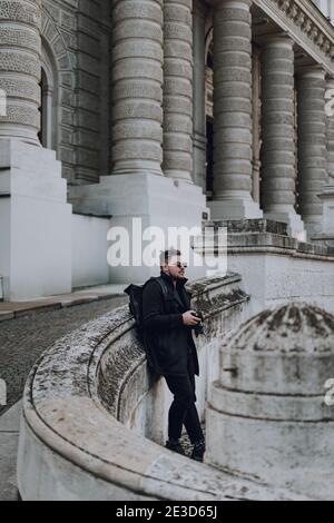 Stilvoller Kerl mit schwarzem Rucksack im Freien stehend und mit Kamera mit Gebäude auf Hintergrund. Reisekonzept Stockfoto