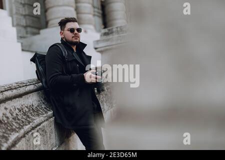 Junger ruhiger Mann mit Rucksack, der Kamera auf der Straße hält, mit Gebäude im Hintergrund. Reisekonzept Stockfoto