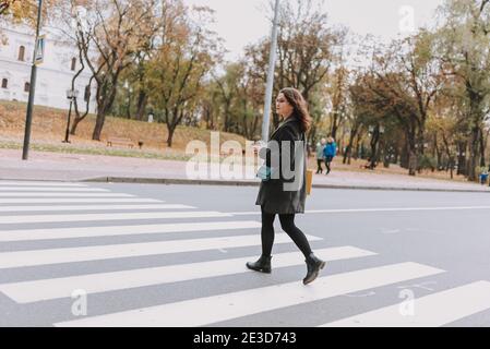 Hübsche junge Frau mit Kaffee zu gehen suchen, um die Links, während Sie die Straße auf einem Fußgängerübergang vorsichtig überqueren Stockfoto