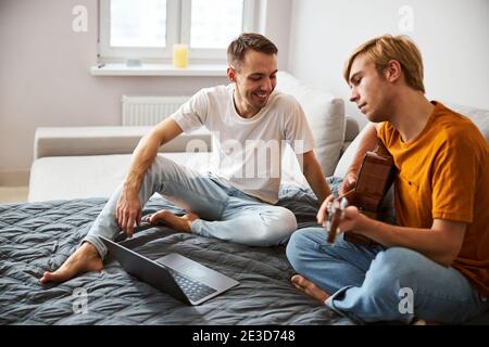 Fröhlicher junger Mann, der seinem Freund zusieht, der Gitarre spielt Stockfoto