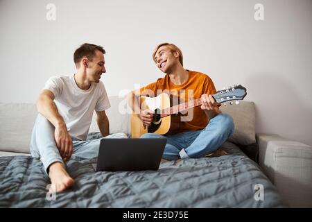 Fröhlicher junger Mann, der seinem Freund zusieht, der Gitarre spielt Stockfoto