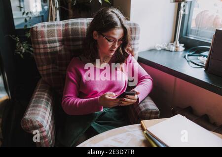 Hübsche Dame mit Brille sitzt im Café beim Betrachen Smartphone-Bildschirm und Eingabe Stockfoto
