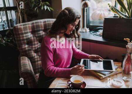 Lächelnde junge Frau mit Brille und Blick auf digitale Tablet Bildschirm beim Sitzen am Tisch im Café Stockfoto