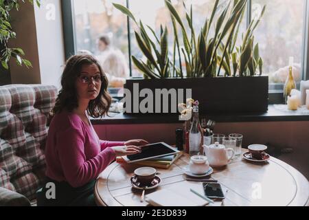 Lächelnde attraktive Frau sitzt im Café während der Arbeit mit digitalen Tablette und Tee genießen Stockfoto