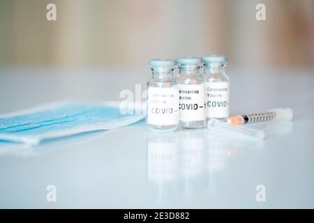 Drei Coronavirus-Impffläschchen, eine Gesichtsmaske und eine Spritze. Stockfoto