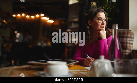 Lächelnde Frau trägt eine Brille und sitzt im Kaffeehaus während Notizen im Notizbuch machen Stockfoto