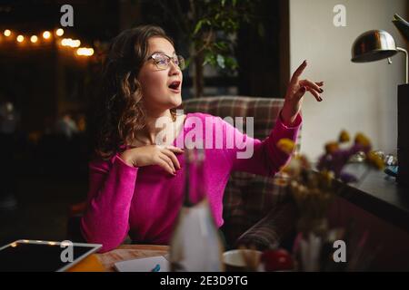 Attraktive junge Dame trägt eine Brille und arbeitet drinnen beim Winken An ihre Freundin vor dem Fenster Stockfoto