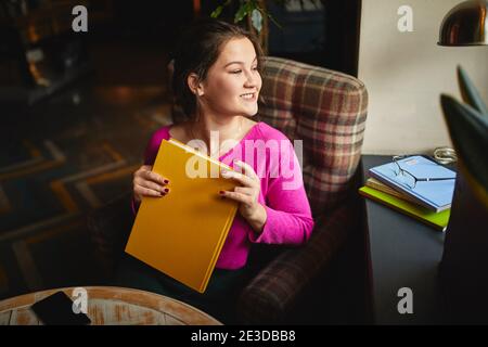 Lächelnde hübsche Dame, die in einem gemütlichen Sessel sitzt und nach draußen schaut Das Fenster in der Cafeteria Stockfoto