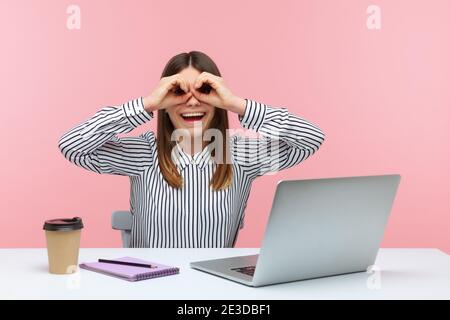 Lächelnd positive Büroangestellte am Arbeitsplatz sitzend, Hände in der Nähe der Augen haltend, vorgebend, in ein Fernglas zu schauen und zu versuchen, jemanden zu finden. Stu Stockfoto