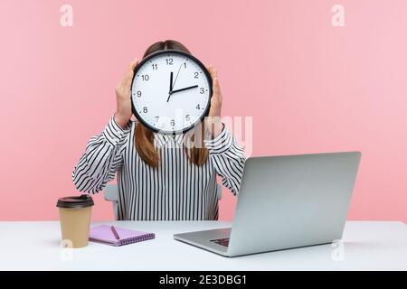 Zeit zum Arbeiten! Frau Büroangestellte sitzt am Arbeitsplatz versteckt Gesicht hinter großen Wanduhr, Zeitmanagement, Zeitplan und Geschäftstreffen Termin. Stockfoto