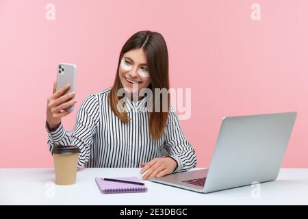 Positive attraktive Frau Büroangestellte Anwendung Flecken unter den Augen und machen Foto auf Smartphone-Kamera sitzen am Arbeitsplatz und lächeln, Spaß haben Stockfoto