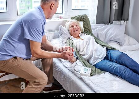Ältere Patientin im Krankenhaus mit besorgten Ehemann, der die Hände hielt, während er den Blutdruck mit Tonometer überprüfte. Mann hilft, Unterstützung Stockfoto