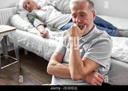 Der ältere Mann sitzt mit ihrer kranken Frau auf dem Bett liegend, fühlt sich schlecht, Frau ist an der Tür des Todes, Mann ist sehr besorgt um sie Stockfoto