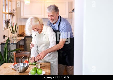 Reife Frau, die Essen zubereitet, ihr Mann hilft ihr von hinten, Gemüse schnitzen Stockfoto