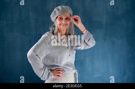 Porträt einer älteren Frau mit grauer Baskenmütze, die vor dunklem Hintergrund im Innenbereich steht. Stockfoto