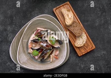 Eine Platte mit Meeresfrüchten, Jakobsmuscheln, Muscheln, Garnelen, Tintenfische, in Sauce, mit Croutons, Brot, auf einem Holzbrett Stockfoto