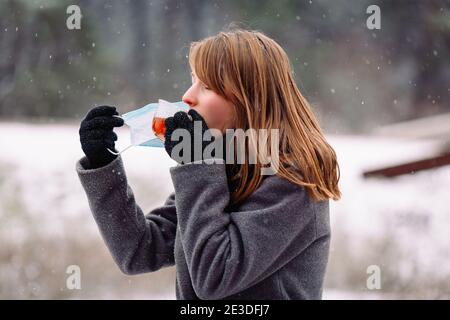 Profilfoto. Kaukasische blonde Mädchen entfernt die medizinische Maske von ihrem Gesicht zur Seite, um den Duft von heißem Tee im Winter gefrorenen Wald zu schmecken. Stockfoto