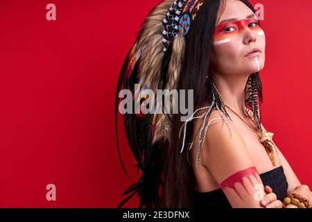 Selbstbewusste indigene Frau in schamanischen tragen Blick auf Kamera isoliert über rotem Hintergrund, mystische indische Frau im Studio. Menschen Individualität, di Stockfoto