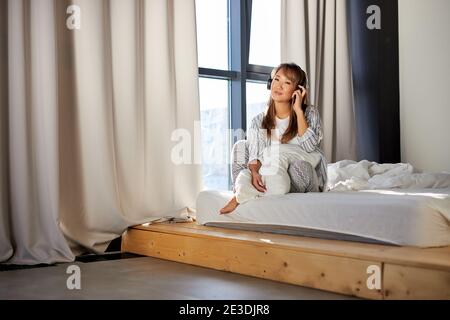asiatische Frau genießt Musik in Kopfhörer sitzen auf dem Bett, glückliche Frau in Pyjamas sitzt allein im Schlafzimmer Stockfoto
