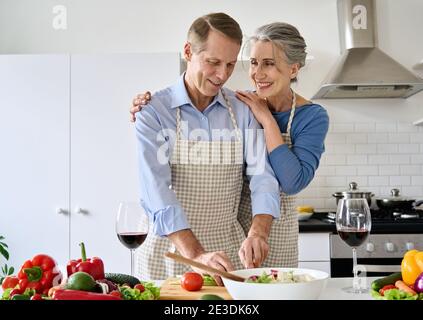 Glückliches altes Paar trinkt Wein Vorbereitung gesunde Ernährung Salat Umarmung zu Hause. Stockfoto